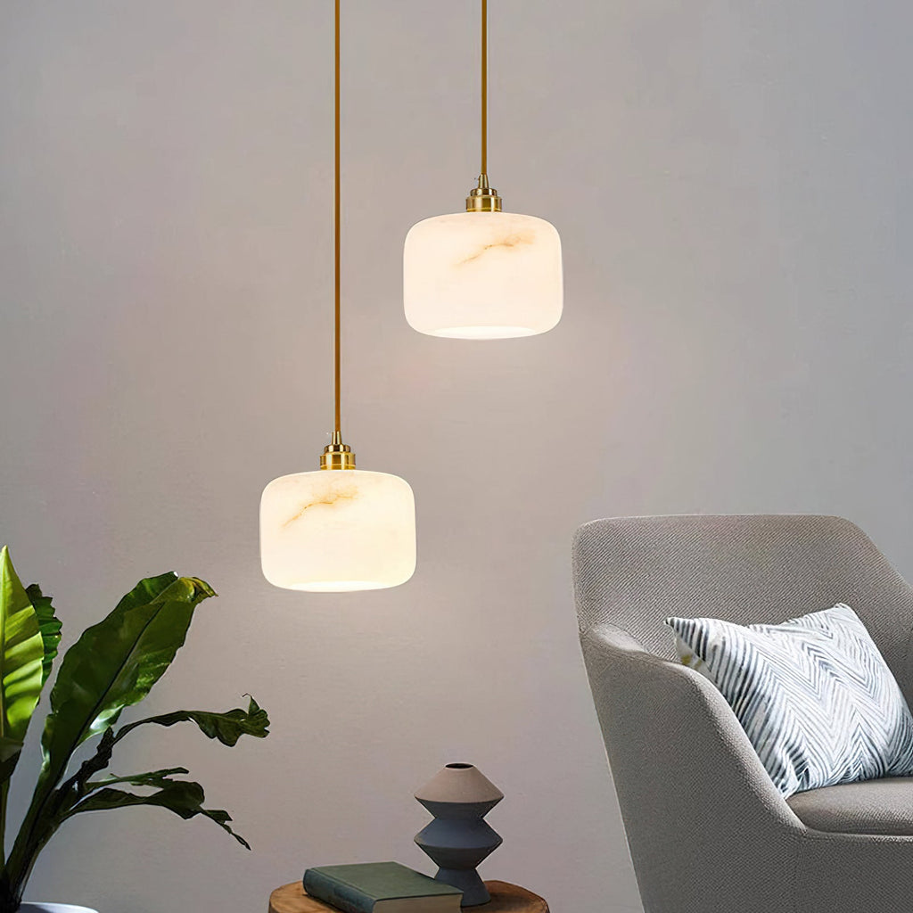 Two Clérachic alabaster pendants with brass fittings hung at different heights beside a grey armchair and a leafy plant.