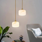 Two Clérachic alabaster pendants with brass fittings hung at different heights beside a grey armchair and a leafy plant.