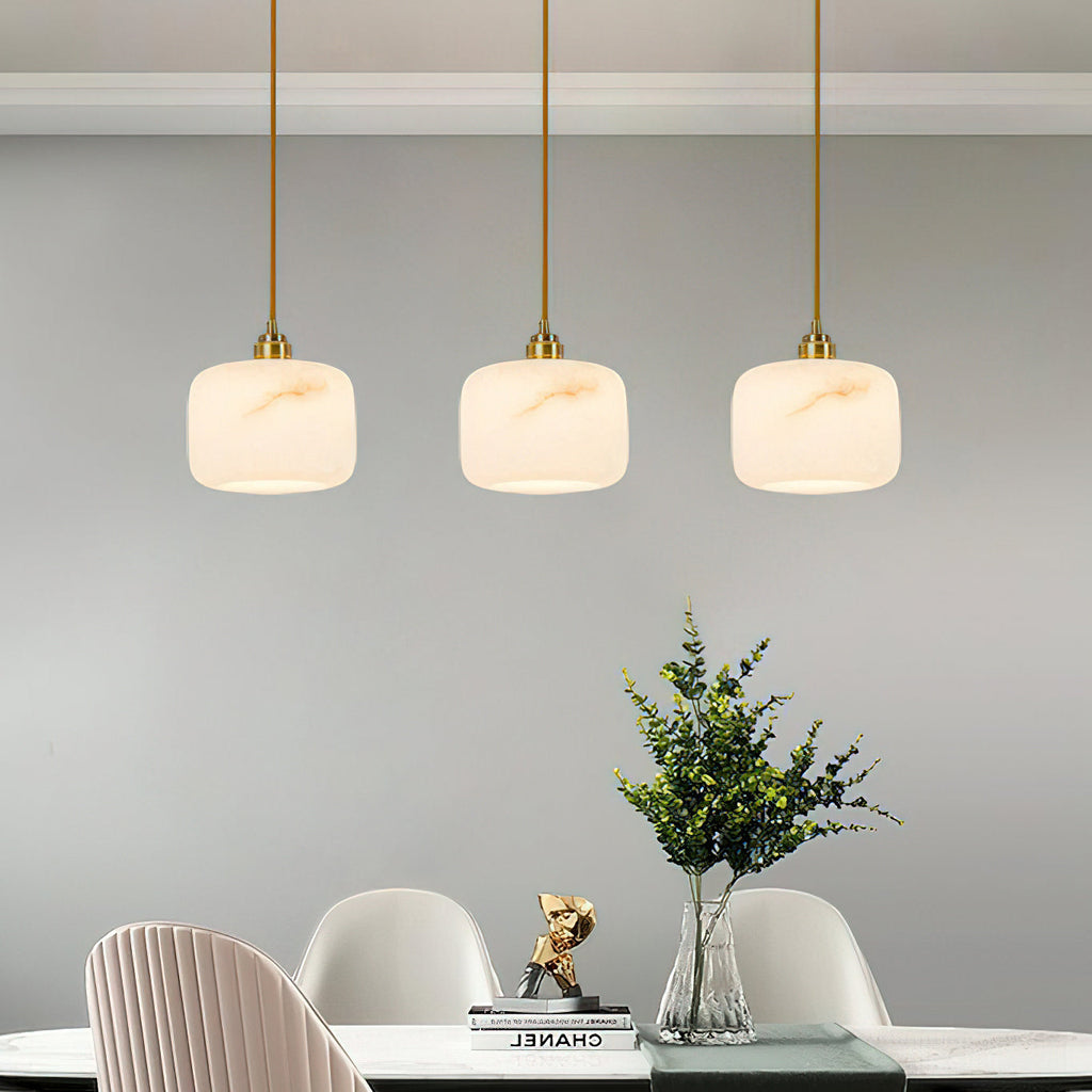 A row of three small Clérachic alabaster pendants over a dining table with pale pink chairs and a green plant.