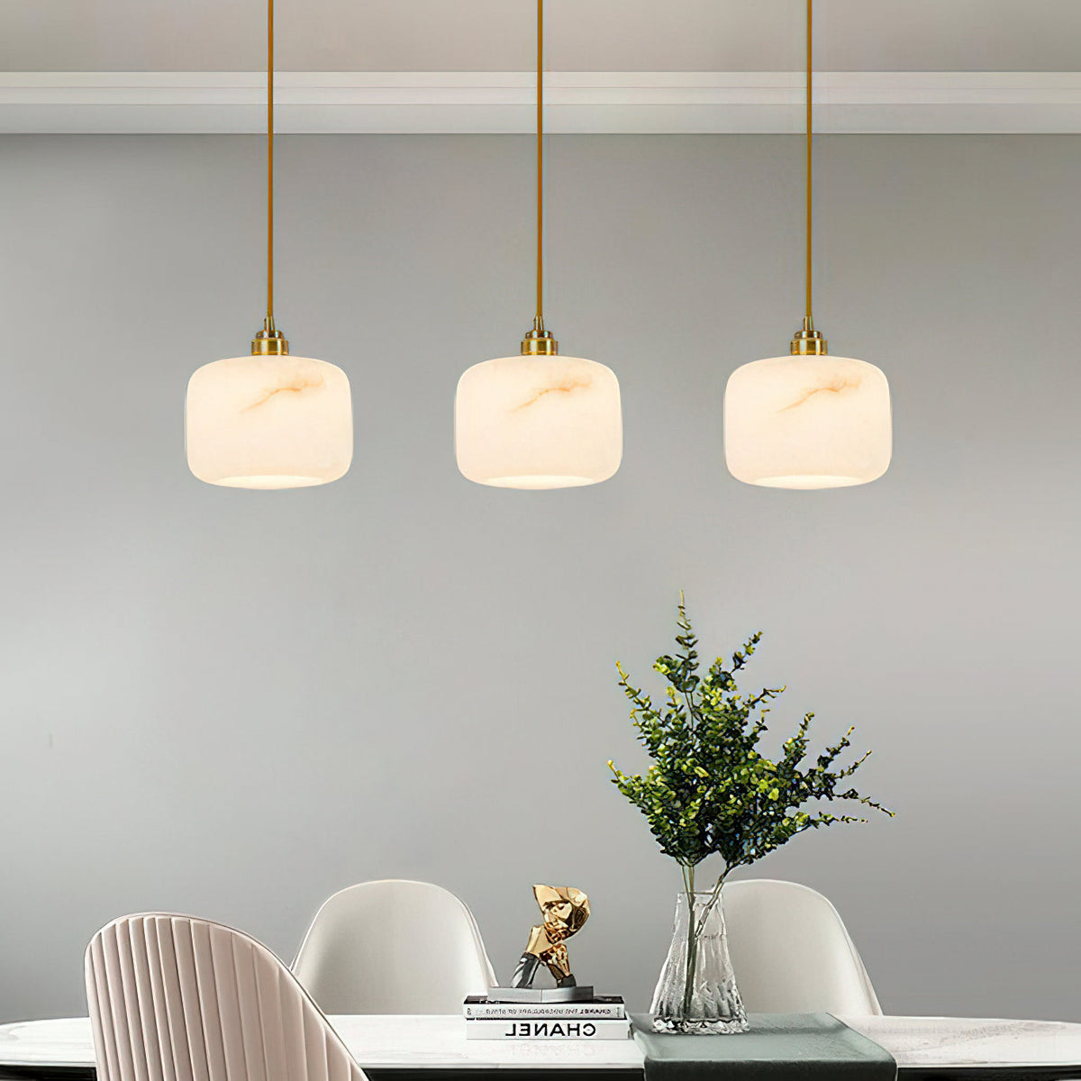 A row of three small Clérachic alabaster pendants over a dining table with pale pink chairs and a green plant.