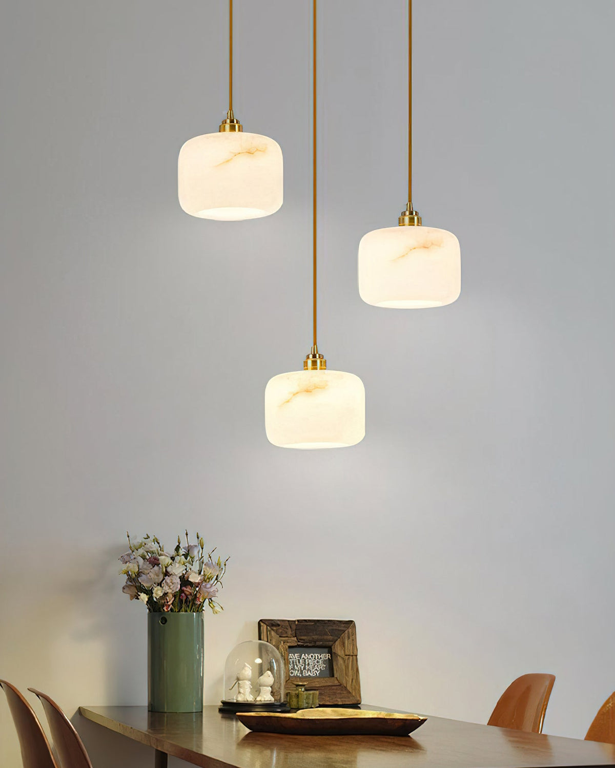 Three Clérachic alabaster pendants hung high above a wooden dining table set with a flower vase and a glass cloche.