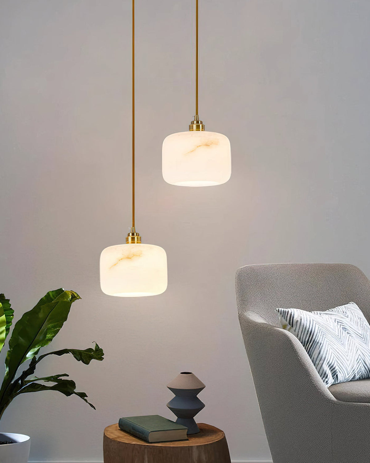 Two Clérachic pendants with brass-capped alabaster shades beside a grey armchair, a leafy plant and a small side table.