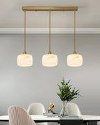 Three Clérachic alabaster pendants hanging in a row from a long brass ceiling bar above a dining table and white chairs.