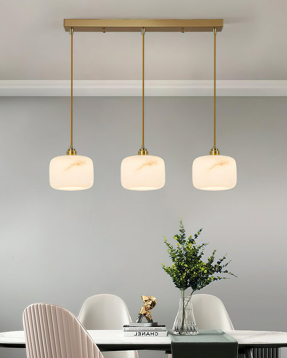 Three Clérachic alabaster pendants hanging in a row from a long brass ceiling bar above a dining table and white chairs.