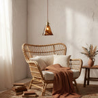 A Clérachic amber glass pendant lit above a rattan armchair with cream cushions and a rust throw, against a rough plaster wall.