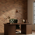 A small Clérachic amber pendant hangs in front of a brick wall, above a walnut desk with a brass desk lamp and a green chair.