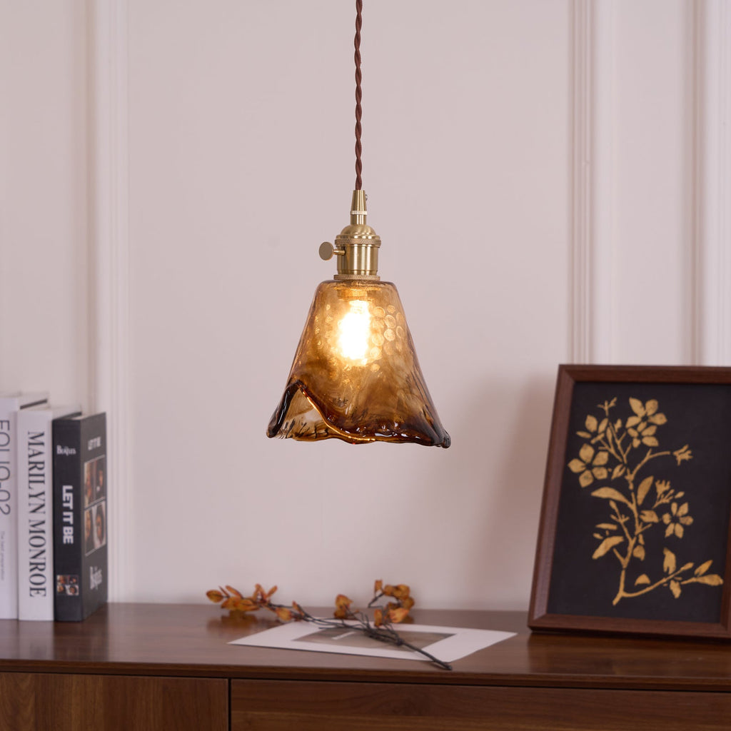 Lit Clérachic pendant with a flared amber glass shade and brass fitting, above a sideboard with books and a gold-on-black framed print.