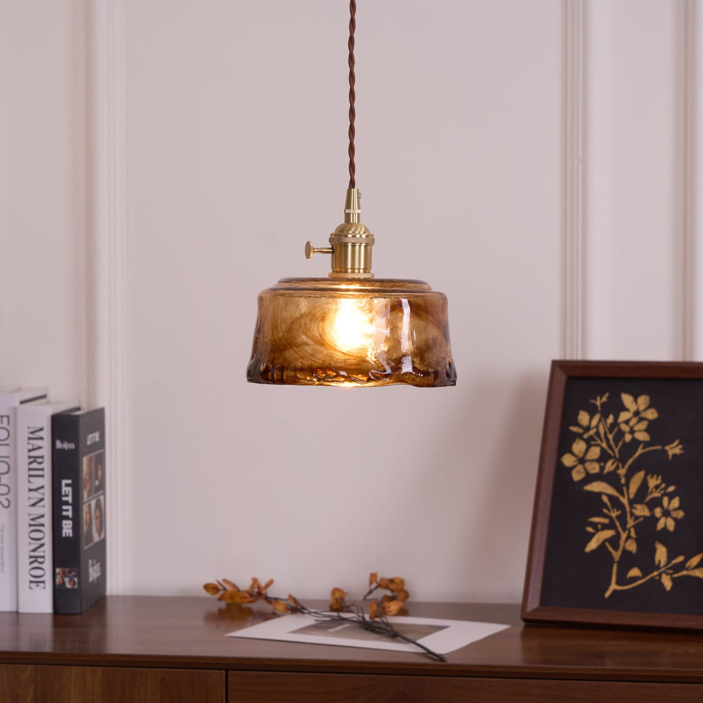 A Clérachic pendant with a low, round amber glass shade, lit above a wooden sideboard with books and a framed botanical print.