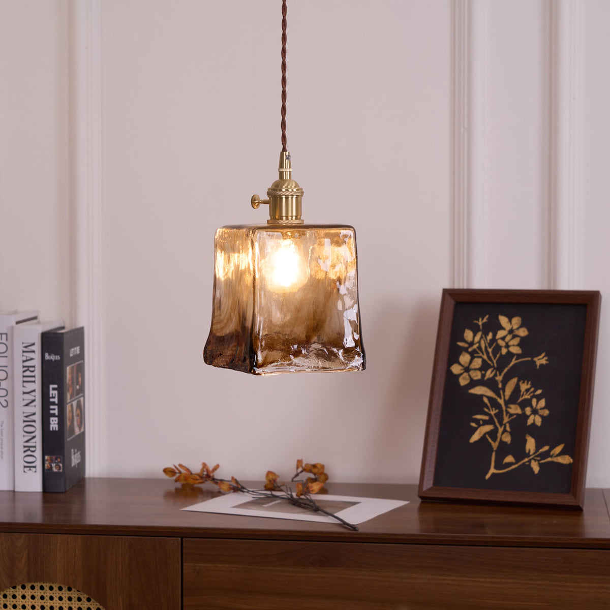 A Clérachic pendant with a squared amber glass shade, lit with a visible bulb, above a walnut sideboard with books and a print.