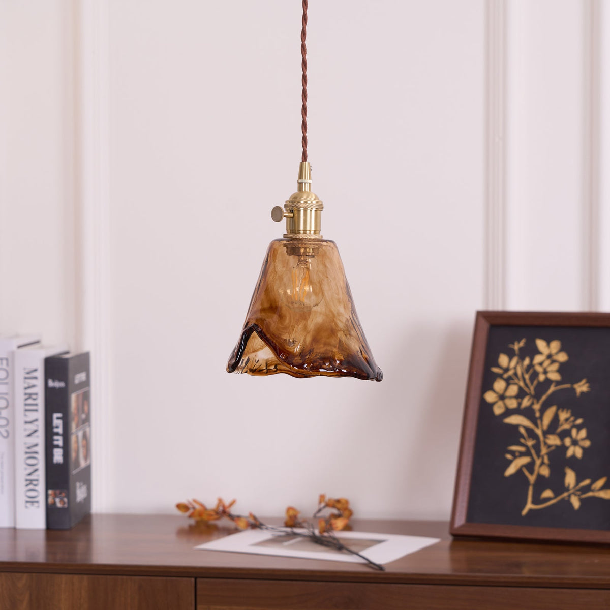 Unlit Clérachic pendant with a flared amber glass shade, with a rippled hem, hanging above a sideboard with a framed print.
