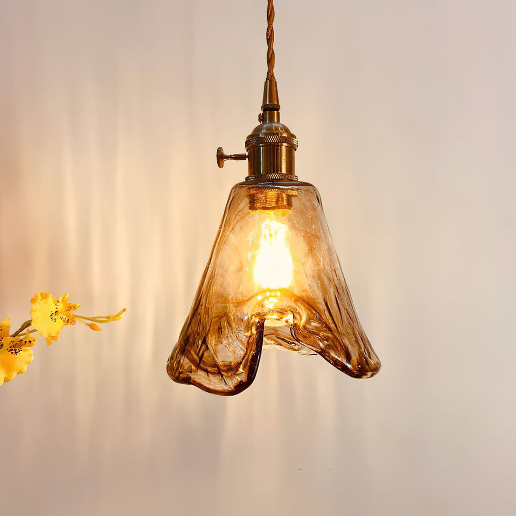Close view of a lit Clérachic pendant with a flared amber glass shade and a brass switch holder, with yellow flowers beside it.