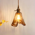 Close view of a lit Clérachic pendant with a flared amber glass shade and a brass switch holder, with yellow flowers beside it.