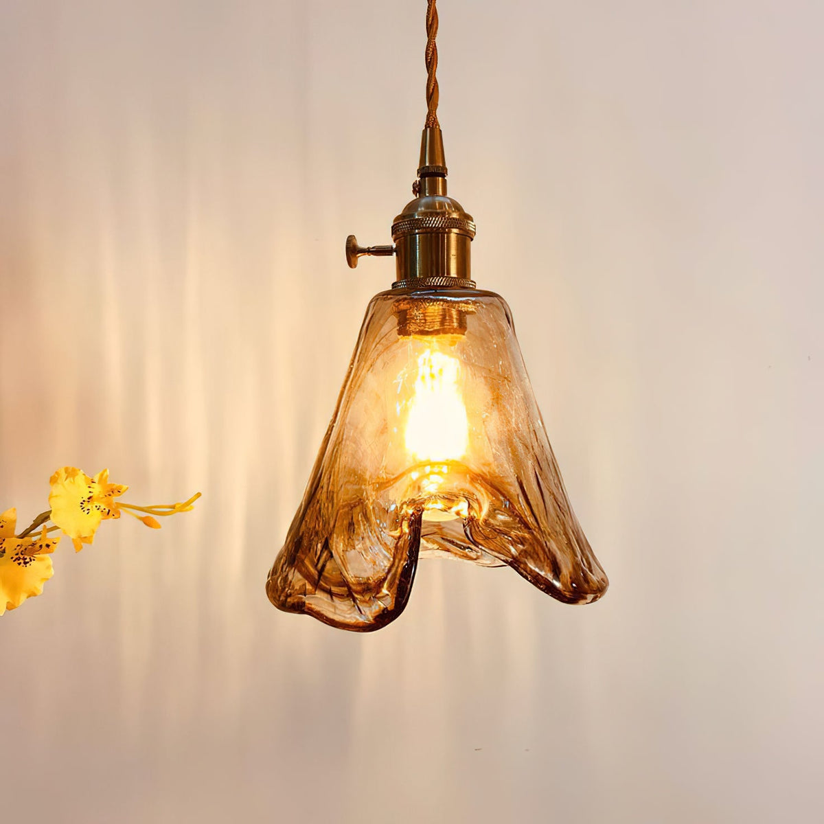 Close view of a lit Clérachic pendant with a flared amber glass shade and a brass switch holder, with yellow flowers beside it.