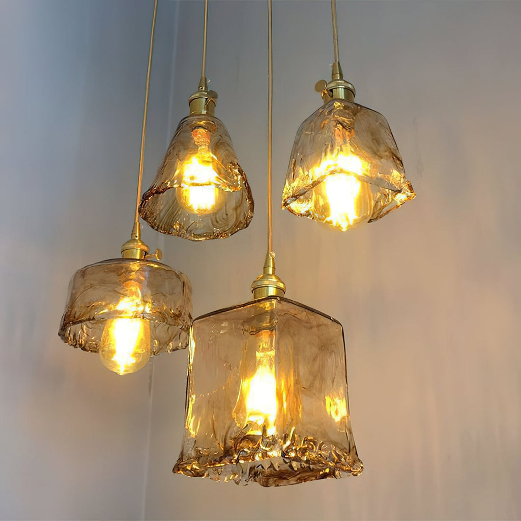 Four lit Clérachic amber glass pendants hang at different heights, in bell, bowl and square shapes, on a blue-grey wall.