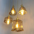 Four lit Clérachic amber glass pendants hang at different heights, in bell, bowl and square shapes, on a blue-grey wall.
