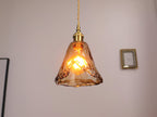 A lit Clérachic pendant with a flared amber glass shade, hanging on a pale wall beside a small gold picture frame.