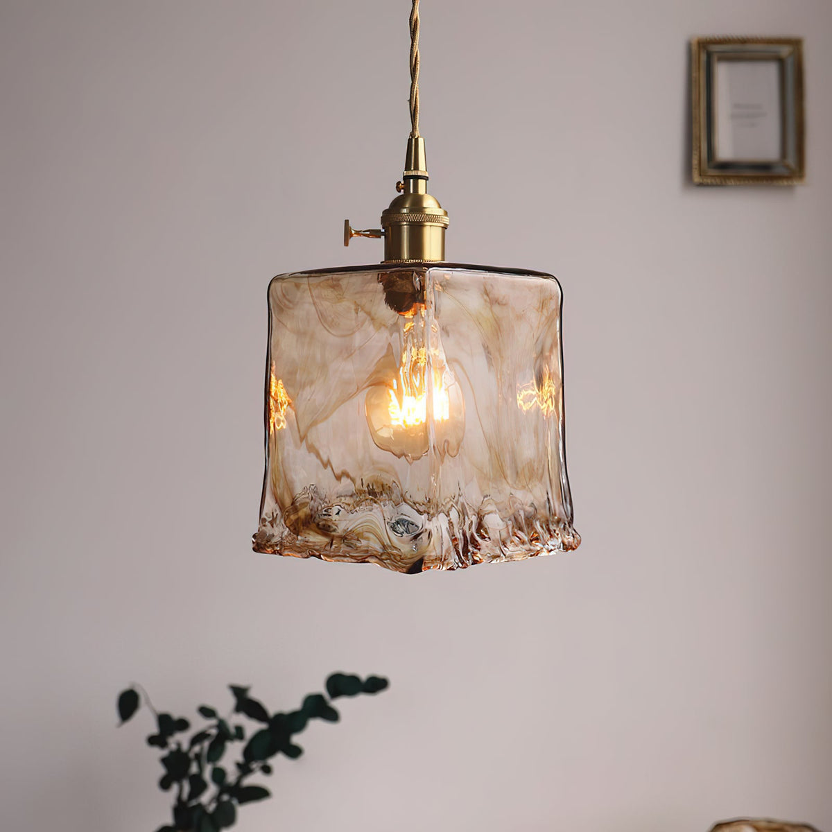 Clérachic pendant with a squared amber glass shade and a visible bulb, hanging by a framed picture, with a plant below.