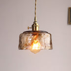 A Clérachic pendant with a low, wide amber glass shade, lit, with a globe bulb showing below the glass, on a pale wall.