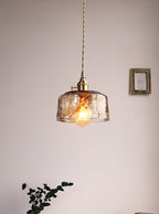 Clérachic pendant with a low, round amber glass shade, seen from a distance with a plant and a small frame nearby.