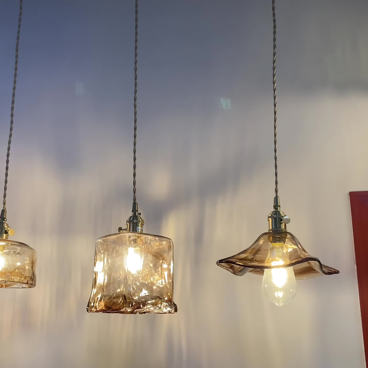 Three Clérachic pendants, one a squared glass, one a bowl and one with a wavy flared rim, lit against a dusk-blue wall.