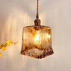 Close view of a lit Clérachic pendant with a squared amber glass shade, with a bunch of yellow flowers at the left.
