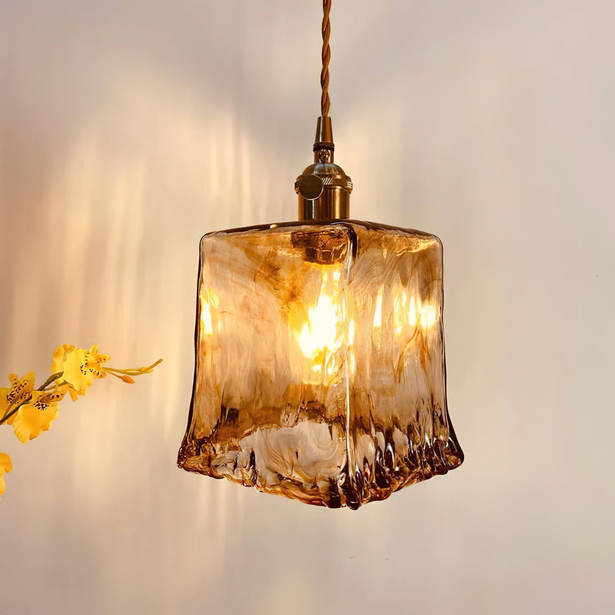 Close view of a lit Clérachic pendant with a squared amber glass shade, with a bunch of yellow flowers at the left.