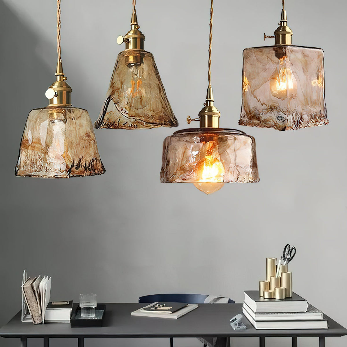 Four Clérachic amber glass pendants hanging at different heights above a dark desk with books, with a grey wall behind.