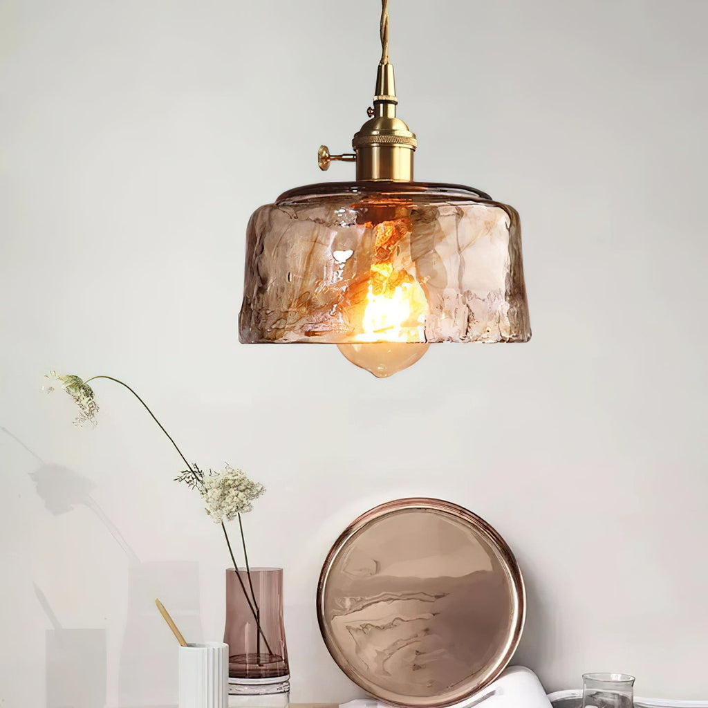 Clérachic pendant with a low, round amber glass shade, lit above a pink glass vase, a copper tray and a stem of white flowers.