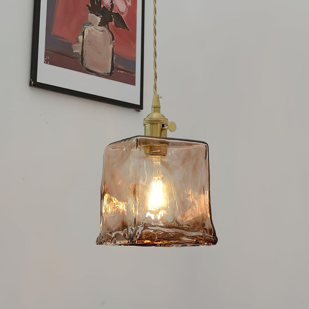 A Clérachic pendant with a squared amber glass shade, lit, hangs below a framed picture, with its brass holder and twisted cord.