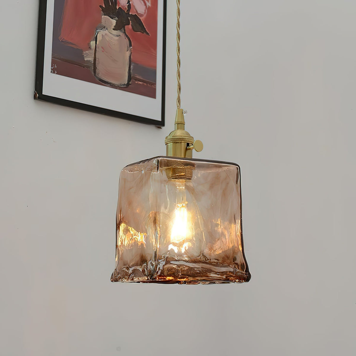 A Clérachic pendant with a squared amber glass shade, lit, hangs below a framed picture, with its brass holder and twisted cord.