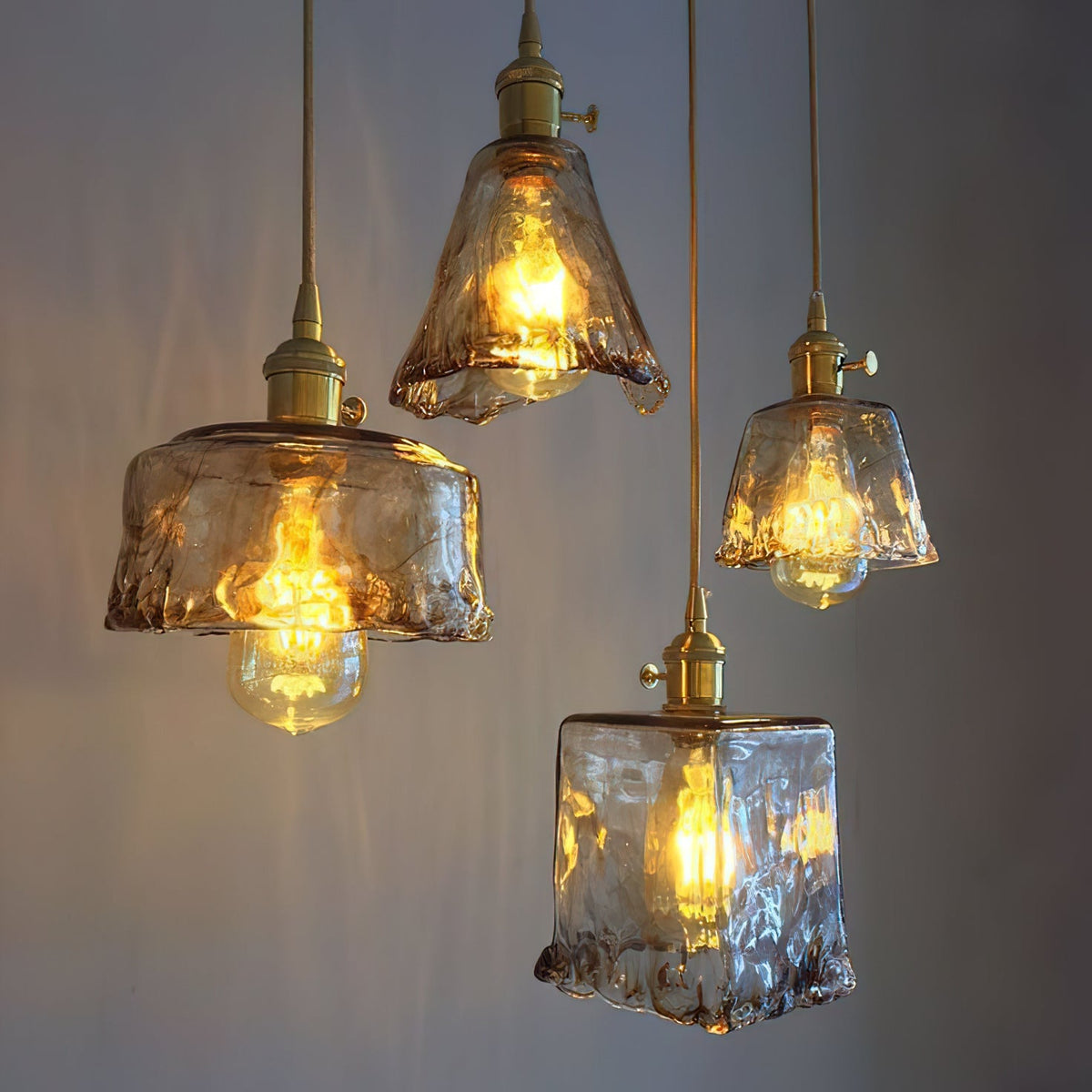 Four Clérachic amber glass pendants, in square, bowl and flared shapes, lit at different heights against a dark wall.