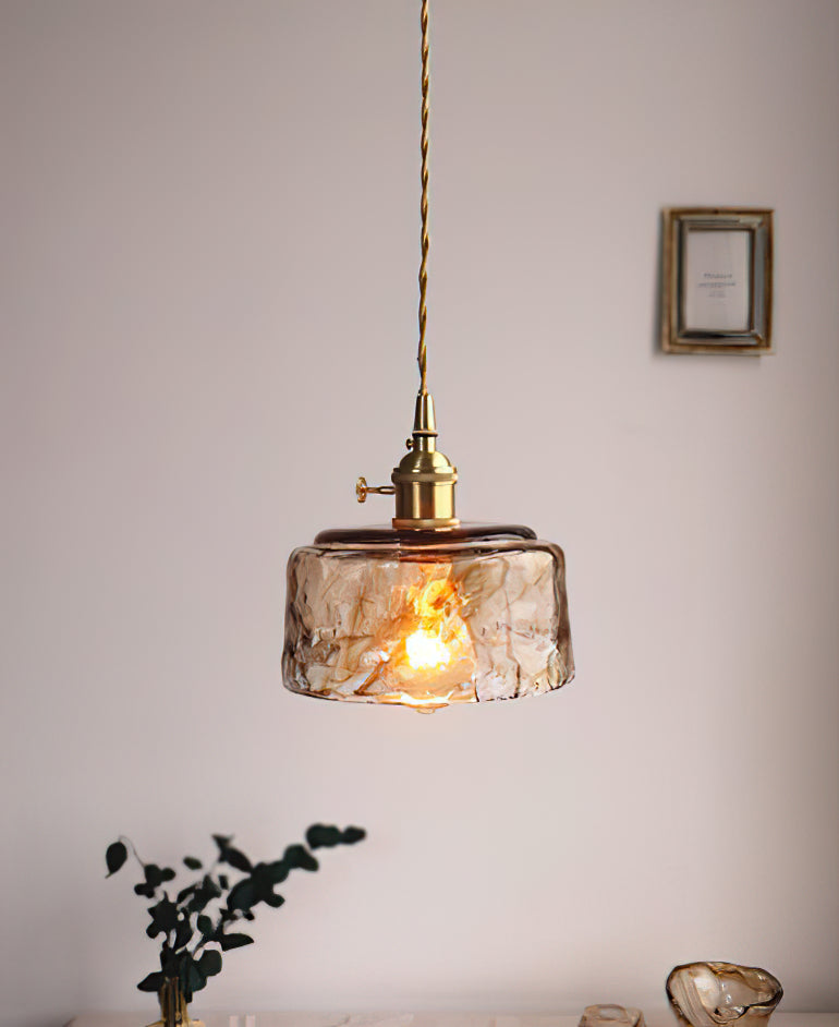 Clérachic pendant with a low, round amber glass shade, lit, hanging by a small frame, with a dark plant below.