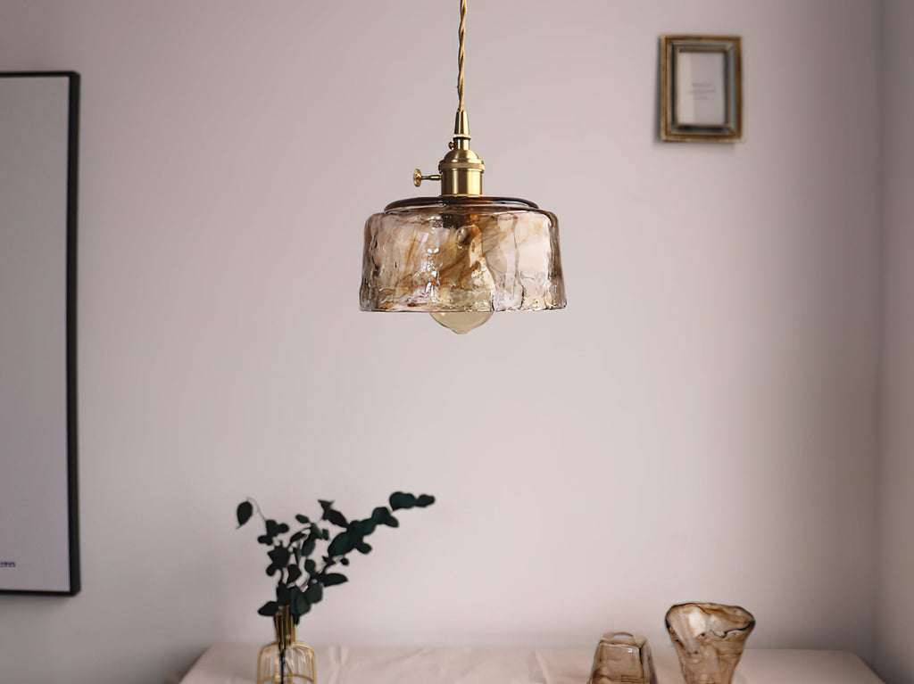 A Clérachic pendant with a low, round amber glass shade, seen from a distance by a dark plant and amber glass pots.