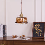 A Clérachic pendant with a low, round amber glass shade, above a sideboard with books, a framed botanical print and dried leaves.
