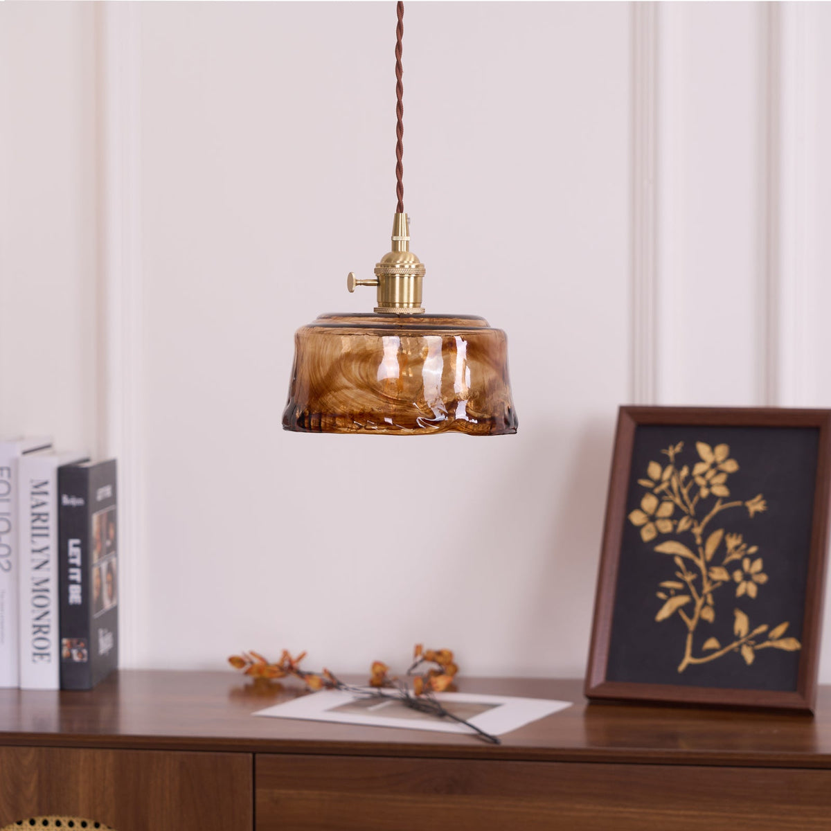 A Clérachic pendant with a low, round amber glass shade, above a sideboard with books, a framed botanical print and dried leaves.