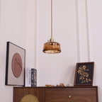 A low, round Clérachic pendant with a brown-and-amber swirled shade hangs above a console with two framed prints and books.