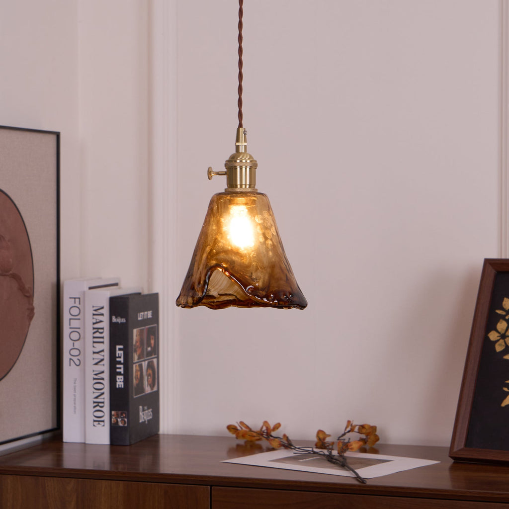 A Clérachic pendant with a flared amber glass shade, lit, hangs beside a book stack and a framed print on a sideboard.