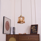 A Clérachic pendant with a squared amber glass shade, unlit, hanging above a cabinet with a cane front and a round print.