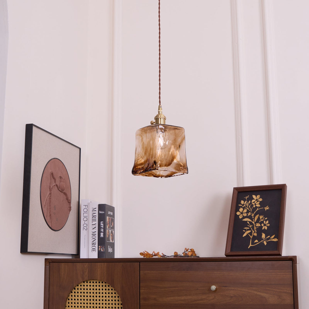 A Clérachic pendant with a squared amber glass shade, unlit, hanging above a cabinet with a cane front and a round print.