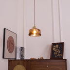 A Clérachic pendant with a low, round amber glass shade, lit, hanging above a cane-front cabinet and a framed round print.