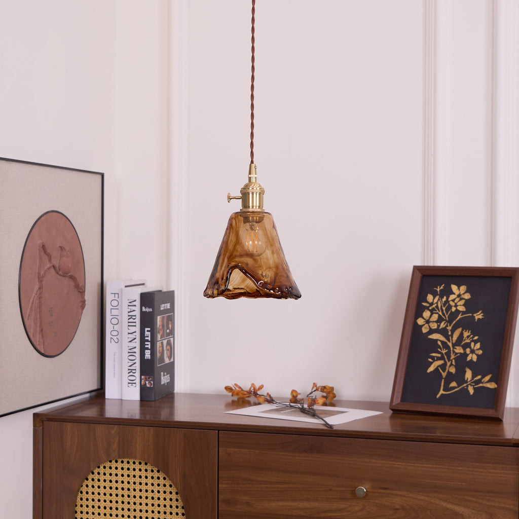 Clérachic pendant with a flared amber glass shade, unlit, hanging above a cabinet with books and a framed botanical print.