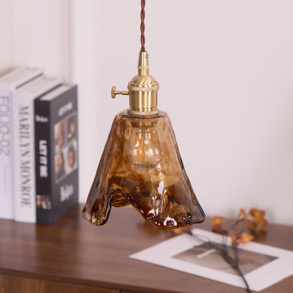 A Clérachic pendant with a flared amber glass shade, lit, with a wavy hem and books on the sideboard below.