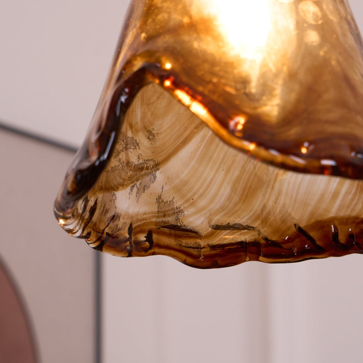 Close view of the wavy, glossy hem of a Clérachic pendant with a flared amber glass shade, lit from within.