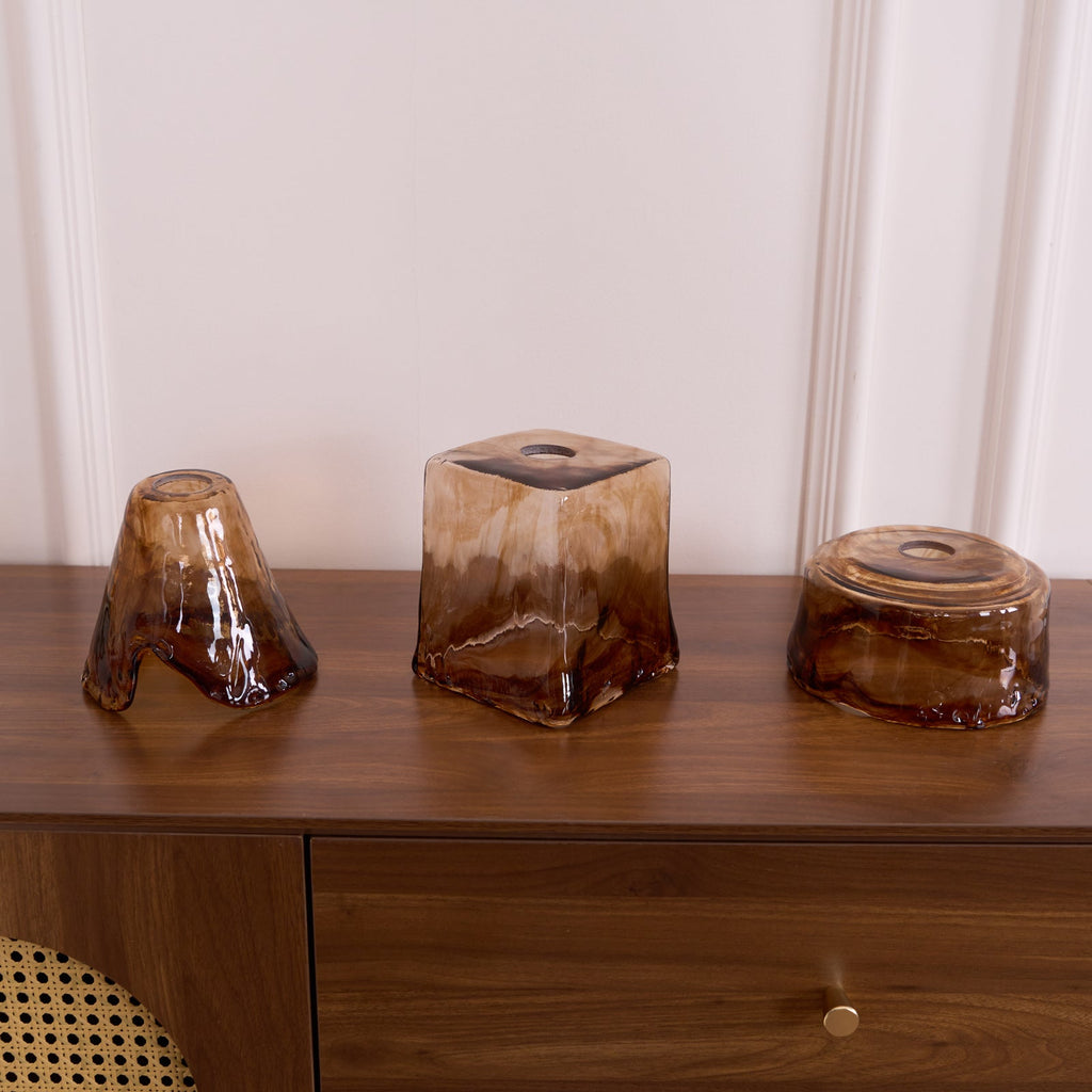Three loose Clérachic amber glass shades on a walnut cabinet, with a flared, a squared and a round shape side by side.