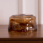 A loose low, round Clérachic amber glass shade on a wooden surface, with a swirled amber and brown pattern and a thick rim.