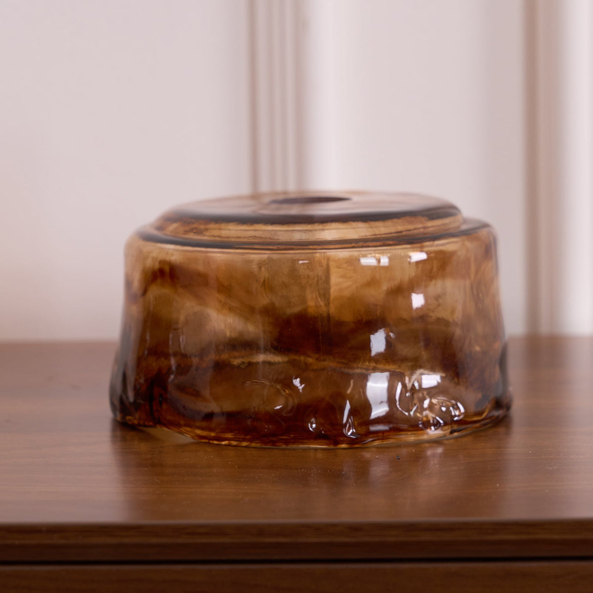 A loose low, round Clérachic amber glass shade on a wooden surface, with a swirled amber and brown pattern and a thick rim.