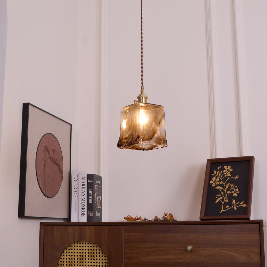A small Clérachic pendant with a squared amber glass shade, lit, hanging above a cabinet, a round print and a framed botanical print.