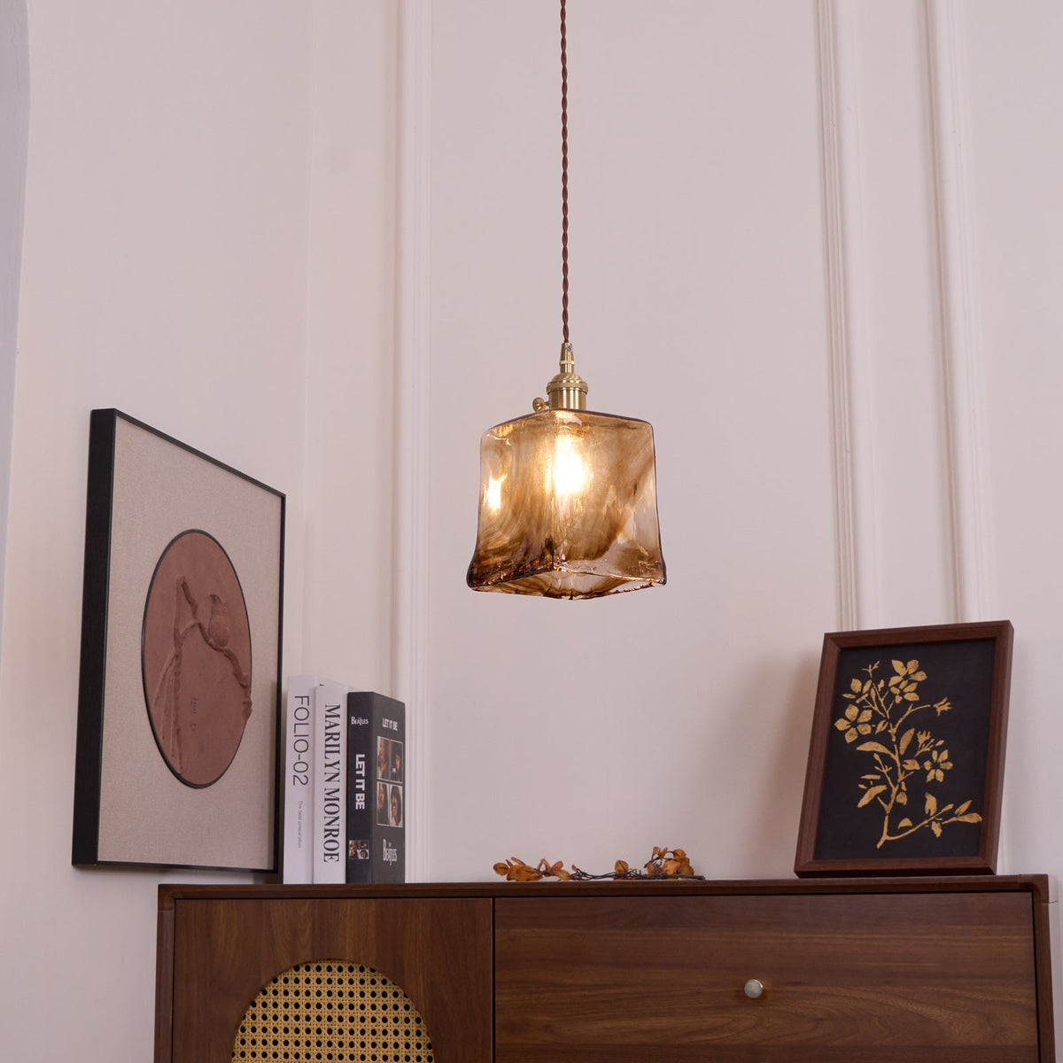 A small Clérachic pendant with a squared amber glass shade, lit, hanging above a cabinet, a round print and a framed botanical print.