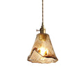 A Clérachic pendant with a flared amber glass shade, lit, on a white backdrop, with a rippled hem and a brass switch holder.