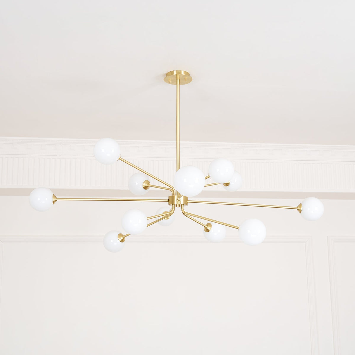 Looking up at a brass Clérachic chandelier with white globes, set against a white ceiling and a moulded cornice.